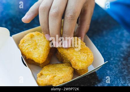 Un enfant atteint une délicieuse portion de pépites frites dorées à partir d'une boîte en papier placée sur la table Banque D'Images