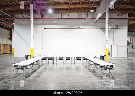 Table et chaises aménagées comme lieu de rencontre dans un grand entrepôt industriel. Sol en béton, murs blancs et austérité. Banque D'Images