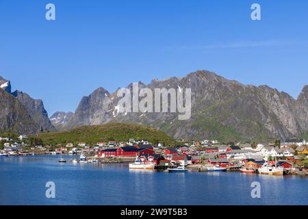 Vue panoramique de Reine avec cabanes de pêche en rorbu rouge classiques, entourée de montagnes majestueuses avec des sommets enneigés dans les îles Lofoten, Norvège Banque D'Images