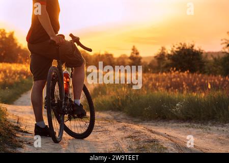 Homme avec vélo debout sur un sentier poussiéreux dans le champ au coucher du soleil. Concept de cyclisme. Banque D'Images