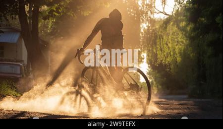 Cycliste faisant du vélo sur une route de gravier au coucher du soleil. Une silhouette de jeune homme sportif sur un vélo de gravier dans un nuage de poussière de vrai well après skiddi Banque D'Images