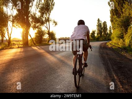 Homme sur un vélo de route roule sur une route asphaltée vide au coucher du soleil. Concept de style de vie actif. Banque D'Images