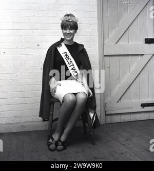 1964, historique, une jeune femme assise sur une chaise en bois dans un coin d'une salle de village avec couronne, ceinture et cape après un concours de beauté et couronnée comme la «Miss Prestwood'. L'ancien village de Prestwood dans les Chilterns, près de Great Missenden dans le Buckinghamshire, Angleterre, Royaume-Uni remonte à l'époque anglo-saxonne. Banque D'Images