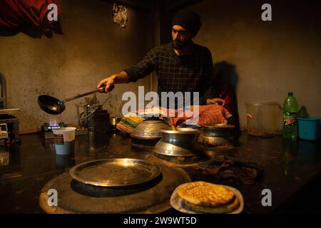 Un chef cachemiri verse de l'huile de friture chaude dans un verre contenant de l'harisa pour servir les clients par une froide matinée à Srinagar. Harisa est un plat de petit déjeuner traditionnel séculaire qui garde le corps au chaud toute la journée pour aider les gens à endurer les conditions de gel. Il est principalement composé de viande de mouton, d'épices, d'aromatiques et de farine de riz. Il faut environ 12 heures pour cuire et doit être stocké pendant la nuit dans une casserole souterraine en terre avec de la vapeur jusqu'à ce que le tout soit écrasé avant la consommation. Ce plat est généralement mangé tôt le matin avec du pain fraîchement préparé. Les historiens pensent que Harisa a été introduit à Kashm Banque D'Images