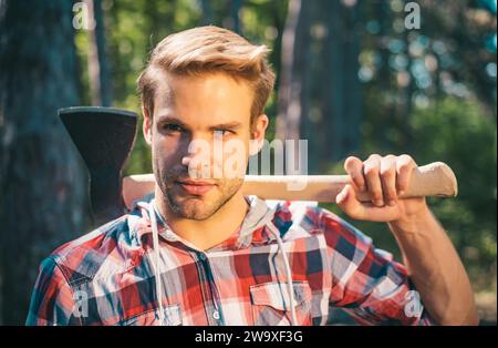 Un beau jeune homme avec une barbe porte un arbre. Beau Woodsworker bûcherons chemise à carreaux tenant la hache sur fond vert nature. Banque D'Images