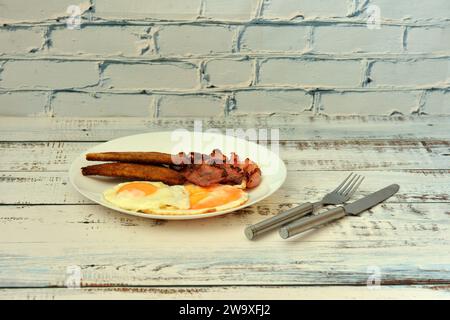 Une assiette avec deux œufs brouillés, des toasts de pain croustillants et des tranches de bacon sur une table en bois clair, couverts à proximité. Gros plan. Banque D'Images