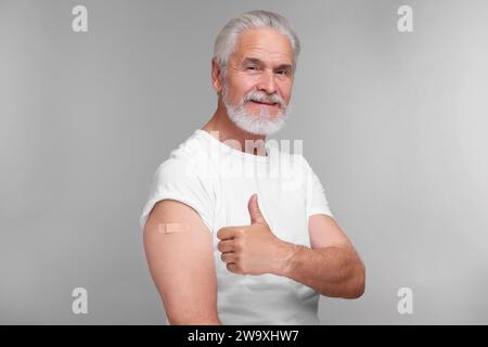 Homme âgé avec un bandage adhésif sur le bras après la vaccination montrant le pouce vers le haut sur fond gris clair Banque D'Images