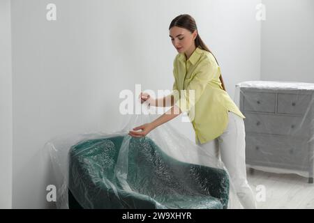 Jeune femme mettant le film plastique loin du fauteuil près du mur gris clair à l'intérieur Banque D'Images