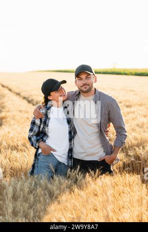 Un couple de fermiers en chemises à carreaux et casquettes se tiennent embrassés sur le champ agricole de blé au coucher du soleil. Banque D'Images