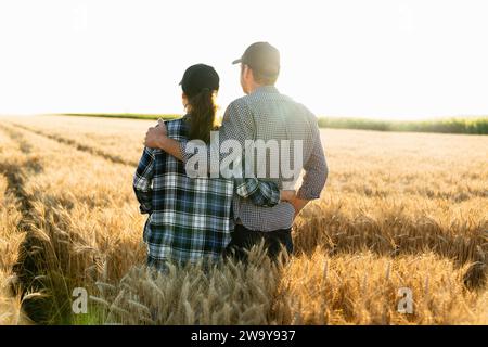 Un couple de fermiers en chemises à carreaux et casquettes se tiennent embrassés sur le champ agricole de blé au coucher du soleil. Banque D'Images