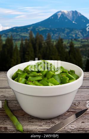 Image minimaliste de nature morte d'un bol en porcelaine blanche contenant des haricots coureurs fraîchement hachés sur une table en bois, dans un paysage avec des arbres et une montagne Banque D'Images