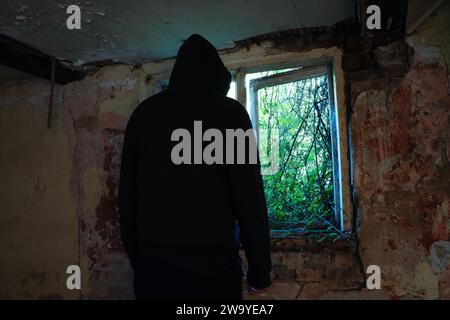 Un concept d'horreur d'une figure effrayante à capuche, de retour à la caméra. Silhouette par une fenêtre dans une maison abandonnée. Avec des murs en décomposition. Banque D'Images