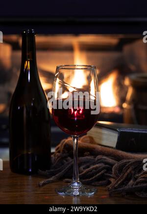Verre de vin rouge avec livre de couverture et bouteille sur la table en bois. Cheminée en feu dans bqckground Banque D'Images
