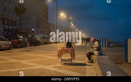 Un Tunisien est assis sur la promenade et regarde son smartphone en attendant de vendre du thé aux passants près de la plage de Bou Jaafar à Sousse, en Tunisie. Banque D'Images