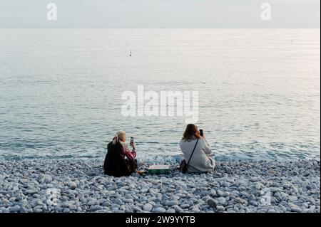 Nice, France. 29 décembre 2023. Deux femmes touristes s'assoient sur la plage de la Promenade des Anglais et regardent le coucher du soleil Banque D'Images