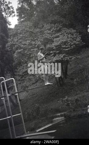 Années 1950, historique, un nageur masculin debout sur un plongeoir, livre de lecture, dans une piscine en plein air boisée, Angleterre, Royaume-Uni. Banque D'Images