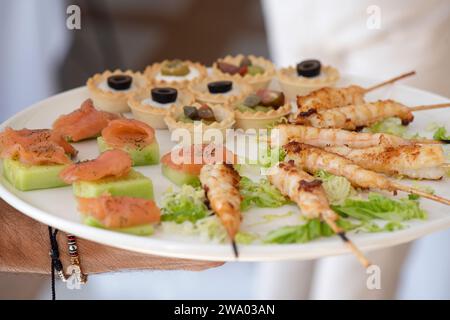 Traiteur tenant une grande assiette avec de petits canapés, une sélection de succulentes brochettes de poulet, des sandwichs au saumon fumé et au concombre et d'autres collations Banque D'Images