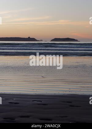 Le coucher du soleil se reflète dans la mer tandis que les vagues roulent vers une plage de sable avec des empreintes de pas de main une île derrière. Essaouira, Maroc. 31 décembre 2023 Banque D'Images