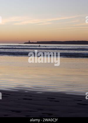 Surfeurs en silhouette en mer avec Island derrière tandis que les vagues roulent vers une plage de sable Essaouira, Maroc. 31 décembre 2023 Banque D'Images