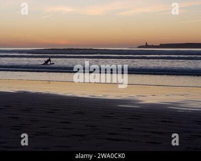 Surfeur en silhouette se trouve sur sa planche au coucher du soleil avec une île derrière et une plage de sable au premier plan. Essaouira, Maroc. 31 décembre 2023 Banque D'Images