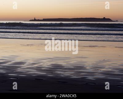 Coucher de soleil sur une île à Essaouira alors que les vagues roulent vers une plage de sable et la lumière orange se reflète dans l'eau. Maroc. 31 décembre 2023 Banque D'Images