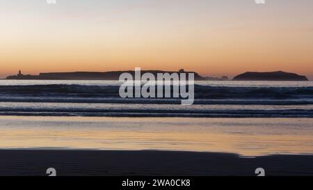 Coucher de soleil sur une île à Essaouira alors que les vagues roulent vers une plage de sable et la lumière orange se reflète dans l'eau. Maroc. 31 décembre 2023 Banque D'Images