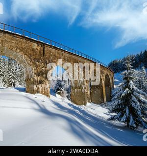 Viaduc de pierre (pont d'arche) sur le chemin de fer à travers la forêt de sapins enneigés de montagne. La neige dévie sur le côté de la voie et le givre sur les arbres et les fils électriques. Banque D'Images