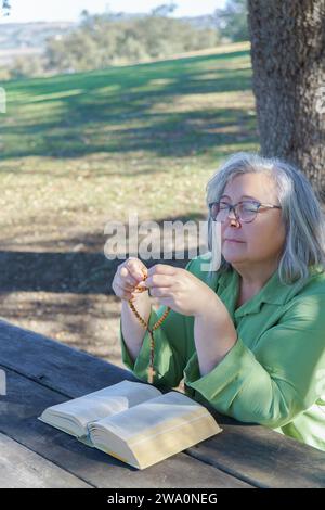 Femme plus âgée aux cheveux blancs avec des perles de chapelet dans la main et une bible priant les yeux fermés assis à une table en bois dans la forêt Banque D'Images