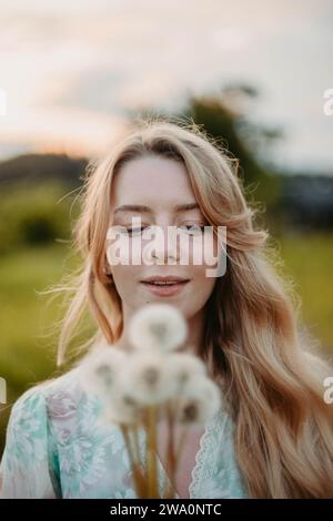 Une belle jeune femme blonde aux cheveux longs dans une robe blanche marche à travers un champ de pissenlits Banque D'Images