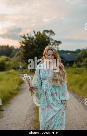 Une belle jeune femme blonde aux cheveux longs dans une robe blanche marche à travers un champ de pissenlits Banque D'Images