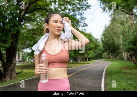 Jeune femme asiatique séduisante essuyant sa sueur de son front pendant sa pause d'eau de sa course d'exercice matinale sur une piste de course d'un local Banque D'Images