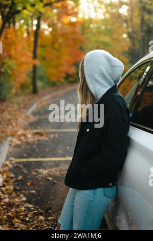 Jeune femme appuyée sur une voiture et regardant les couleurs d'automne dans le parking Banque D'Images