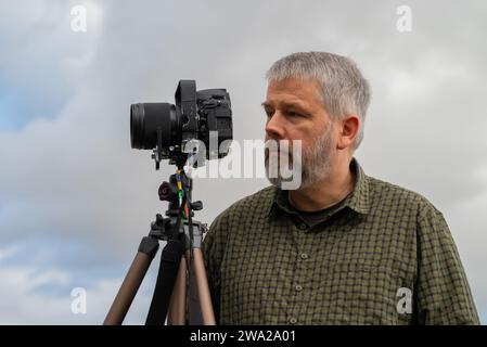 Photographe au travail, cheveux gris et barbe complète, 56 ans, semble réfléchi, chemise à carreaux vert olive, appareil photo reflex 35mm sur trépied, ciel couvert gris Banque D'Images