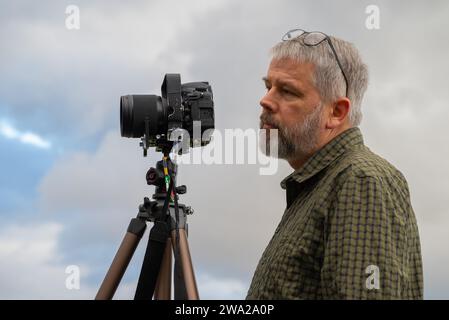 Photographe au travail, cheveux gris et barbe complète, 56 ans, regardant devant l'appareil vers le sujet, lunettes poussées, chemise à carreaux vert olive, Banque D'Images