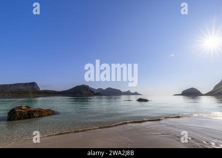 Soleil scintillant le matin sur la paisible plage de Haukland en Norvège, avec des eaux claires et des îlots rocheux sur fond de montagne. Île Lofoten Banque D'Images