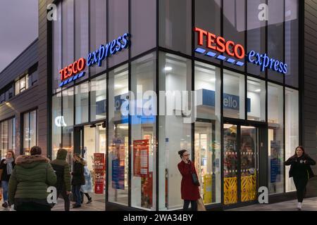 Acheteurs marchant dans et hors d'un Tesco Express au crépuscule dans les centres commerciaux, Basingstoke, Royaume-Uni. Thème - coût de la vie, inflation alimentaire, coût des achats Banque D'Images