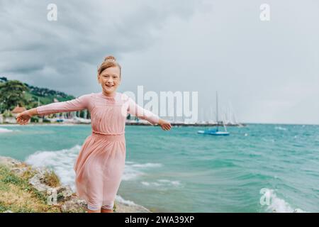 Douce petite fille jouant au bord du lac sur une journée très venteuse portant une robe rose. Image prise sur le lac Léman, Lausanne, Suisse Banque D'Images