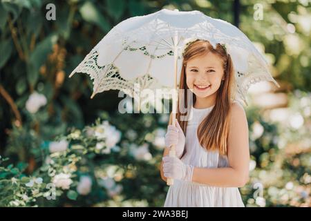 Portrait extérieur de petite fille romantique, portant robe blanche, gants, bandeau de fleur, tenant parapluie de dentelle Banque D'Images
