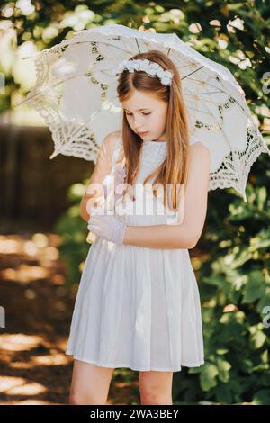 Portrait extérieur de petite fille romantique, portant robe blanche, gants, bandeau de fleur, tenant parapluie de dentelle Banque D'Images