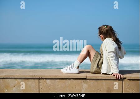 Portrait en longueur d'une adorable petite fille caucasienne assise sur le bord de la mer, respirant l'air frais et profitant d'une belle vue sur les vagues poundin Banque D'Images