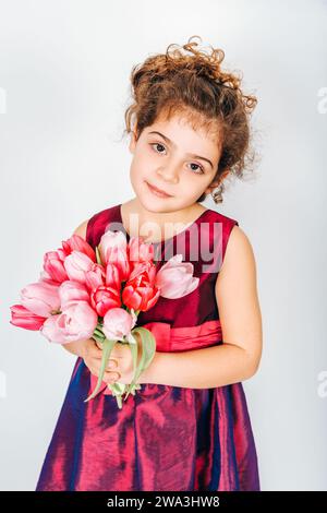 Photo en studio d'une petite fille de 5 ans aux cheveux bouclés, portant une robe de fête, tenant un petit bouquet de tulipes roses fraîches Banque D'Images