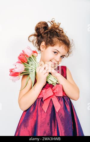 Photo en studio d'une petite fille de 5 ans aux cheveux bouclés, portant une robe de fête, tenant un petit bouquet de tulipes roses fraîches Banque D'Images