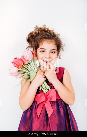Photo en studio d'une petite fille de 5 ans aux cheveux bouclés, portant une robe de fête, tenant un petit bouquet de tulipes roses fraîches Banque D'Images