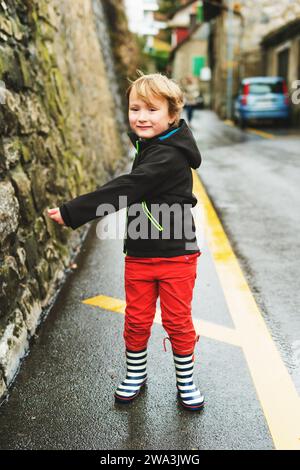 Petit garçon jouant à l'extérieur au début du printemps, marchant à côté de la route, portant une veste imperméable noire, un pantalon rouge et des bottes de pluie Banque D'Images