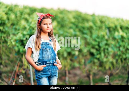 Portrait en plein air de jolie petite fille portant des combinaisons en denim et un bandeau vintage rouge, les mains dans les poches Banque D'Images