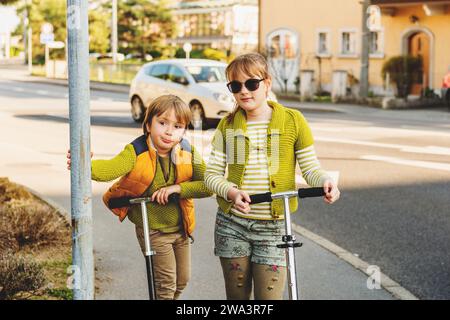 Deux enfants heureux jouant à l'extérieur, garçon de mode et fille posant dans la rue, chevauchant des scooters à côté de la route Banque D'Images
