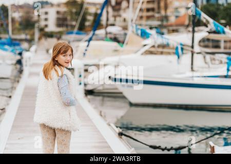 Portrait de mode en plein air de jeune fille préadolescente portant une casquette blanche, gilet en fausse fourrure, jouant dans un port, image tonique Banque D'Images