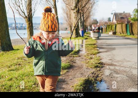 Mignon petit garçon de 3 ans jouant à l'extérieur le jour ensoleillé de début de printemps, portant une veste verte et un chapeau orange Banque D'Images