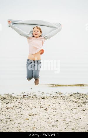 Adorable petite fille de 8-9 ans jouant au bord du lac, portant un pantalon gris et un long cardigan, sautant en l'air Banque D'Images