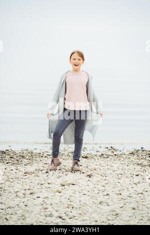 Adorable petite fille de 8-9 ans jouant au bord du lac, portant un pantalon gris et un long cardigan Banque D'Images